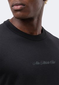 Κοντινό πλάνο στον λαιμό και το πάνω μέρος του στήθους ενός ατόμου που φοράει μαύρο υφασμάτινο πουκάμισο με το κείμενο «Nike Athletic Club» στο μπροστινό μέρος.