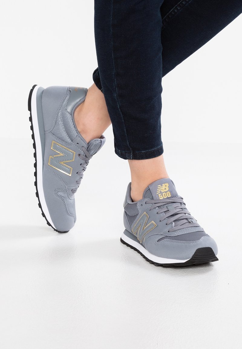 Personne portant des baskets New Balance 500 grises avec des accents dorés et un pantalon noir, debout sur un sol blanc.