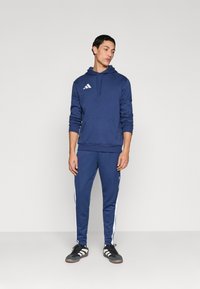 Ναυτικό μπλουζάκι με κουκούλα και τσέπη μπροστά, με λογότυπο τριών λευκών γραμμών· παντελόνι που ταιριάζει με λευκές πλευρικές ρίγες· φοριέται με μαύρα αθλητικά παπούτσια.