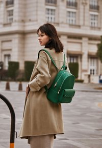 Green leather backpack with a smooth texture, featuring zippers and a front pocket, worn over a beige coat.