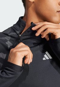 Zwarte sportieve zip-up jas van rekbare stof, met een hoge kraag, reflecterende accenten en een logo op de borst.