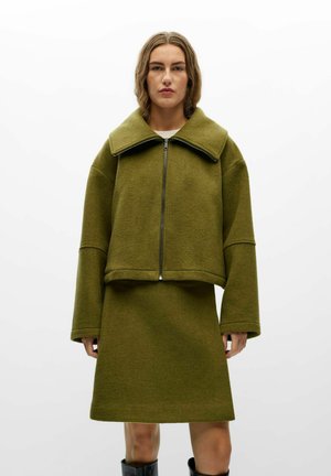 Modèle portant une veste courte vert olive à col large et fermeture éclair sur le devant, assortie à une jupe au genou et à des bottes noires.