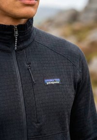 Férfi fekete Patagonia cipzáras polárdzsekiben szabadban, bemutatva a mellkas területét és a hegyeket ábrázoló, szöveges logófoltot.