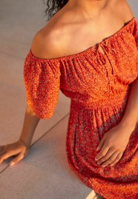 Robe rouge décolletée avec des manches bouffantes, motifs floraux et taille smockée. Modèle assise sur une surface lisse, la main posée à proximité.