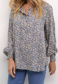 Haut boutonné crème et marine avec un motif de feuilles. Caractéristiques : manches longues avec poignets froncés et encolure sans col.