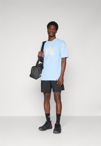 Šviesiai mėlynas marškinėliai su spalvingais piešiniais, derinami su juodomis šortais ir juodais sportba�čiais. Modelis laiko mažą juodą rankinę.