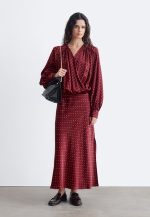 Femme aux longs cheveux foncés portant un chemisier et une jupe à pois bordeaux, tenant un sac à main noir avec une chaîne dorée.