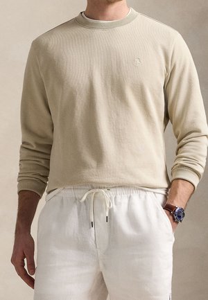Homme portant un sweat-shirt beige à manches longues et un pantalon blanc à cordon de serrage, avec une montre bleu foncé au poignet gauche, debout devant un fond neutre.