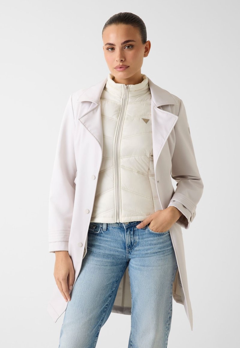 Femme portant un trench-coat beige clair par-dessus une veste matelassée blanche zippée et un jean bleu clair, debout avec une main dans la poche.