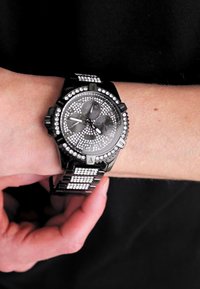 Svart kronografklocka med en strukturerad grå urtavla, silverdetaljer och en armband med diamantliknande dekorationer.