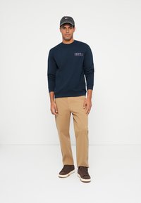 Mornarski pulover s logotipom, kaki hlače in rjavi športni copati. Kapa z logotipom dopolnjuje obleko. Sproščen dizajn, udoben kroj.