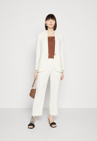 Bílý tailored blazer a široké kalhoty v kombinaci s hnědým saténovým topem. Černé pletené ploché sandály a malá hnědá kabelka doplňují vzhled.