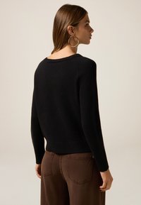 Suéter negro de punto con cuello redondo y dobladillo acanalado, combinado con pantalones marrones. La tela parece suave con una textura acogedora.