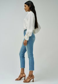 Blusa blanca texturizada, jeans de tiro alto azul claro y tacones marrón claro con tiras. Largo cabello oscuro cae en cascada, visto desde un perfil lateral.