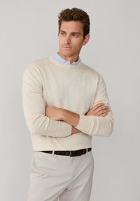 Pull beige à col rond, tissu texturé, porté avec une chemise à col bleu clair en dessous. Pantalon blanc et ceinture sombre.