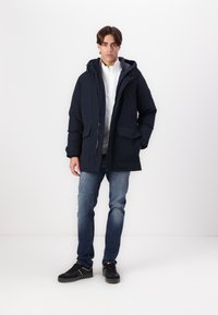 Parka blu navy imbottita con cappuco, zip frontale e due grandi tasche, indossata sopra una camicia bianca e abbinata a jeans e sneakers.