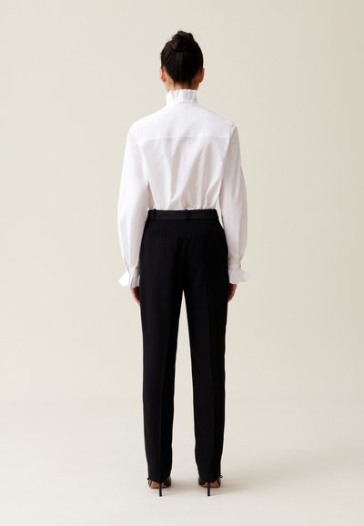 Camicia bianca a maniche lunghe con colletto e polsini arricciati, abbinata a pantaloni neri su misura. Tessuto liscio con una vestibilità strutturata.