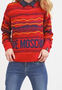 Červený pletený svetr s vodorovnými vzory ve žluté a tmavě modré barvě. Velký tmavě modrý nápis "LOVE MOSCHINO" v oblasti pasu. Žebrované manžety a lem.