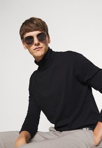 Suéter negro de cuello alto, pantalones grises y gafas de sol redondas con montura de carey. Fondo sencillo, postura sentada.