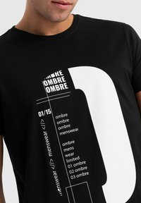 Musta puuvillapaita, jossa on rohkeat valkoiset graafiset printit, mukaan lukien teksti "OMBRE" ja suuri numero "0" suunnittelu etupuolella.