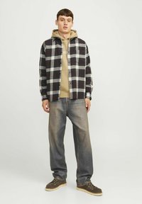 Sort og hvidternet skjorte over en beige hættetrøje, parret med blå denimbukser og mørke sko med en lys sål. Frontvinkel af modellen.