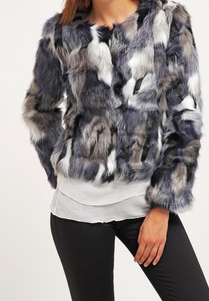 Femme portant une veste en fausse fourrure multicolore grise, noire et blanche par-dessus un chemisier blanc et un pantalon noir.