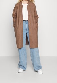 Person iført lang brun frakke, lyserød Levi's t-shirt, brede lyseblå jeans med sort bælte og hvide sneakers, stående mod hvid baggrund.