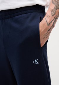 Pantalones de chándal azul marino hechos de algodón suave, con una cinturilla elástica, bolsillos laterales y un logo bordado en azul claro en la pierna izquierda.