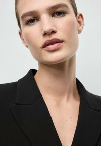 Zwarte blazer met een gestructureerde kraag, soepel materiaal en een aansluitend ontwerp, gedragen door een model met een neutrale make-up en een subtiele oorbellen.