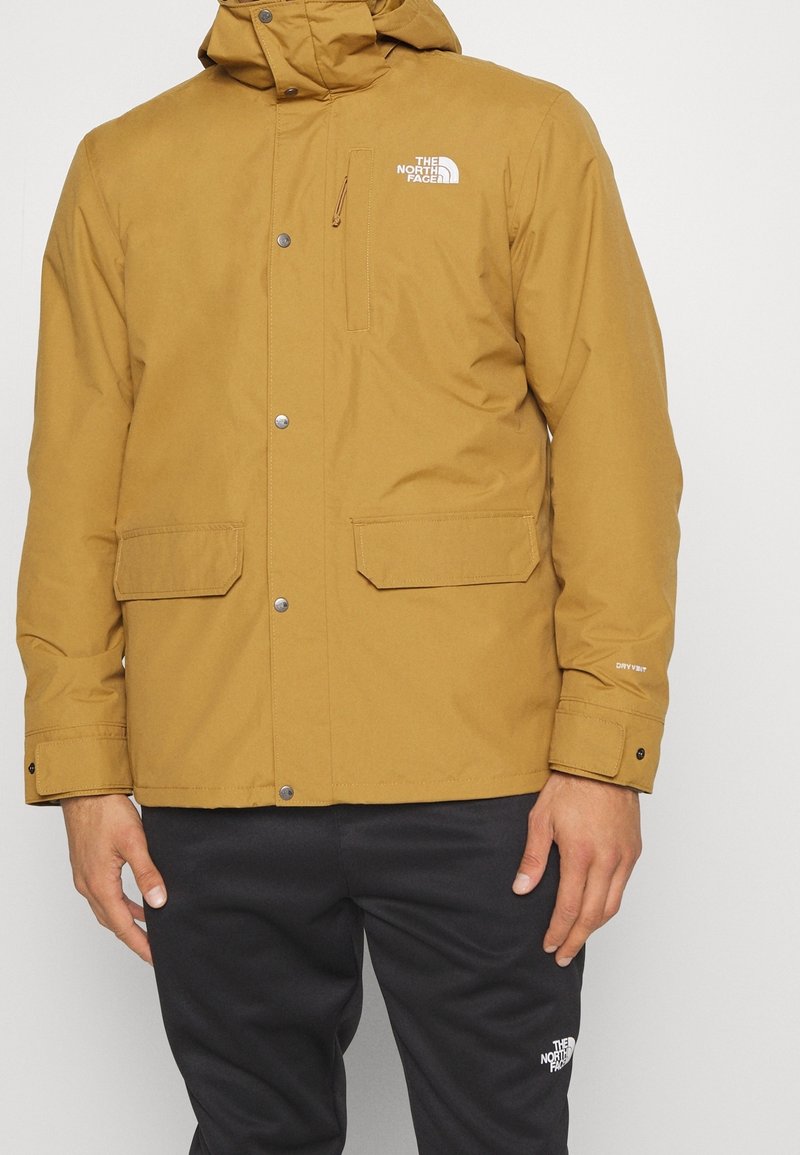 Herrjacka i senapsgul, vattentät med huva, samt svarta byxor, båda med The North Face-logotypen i vitt.