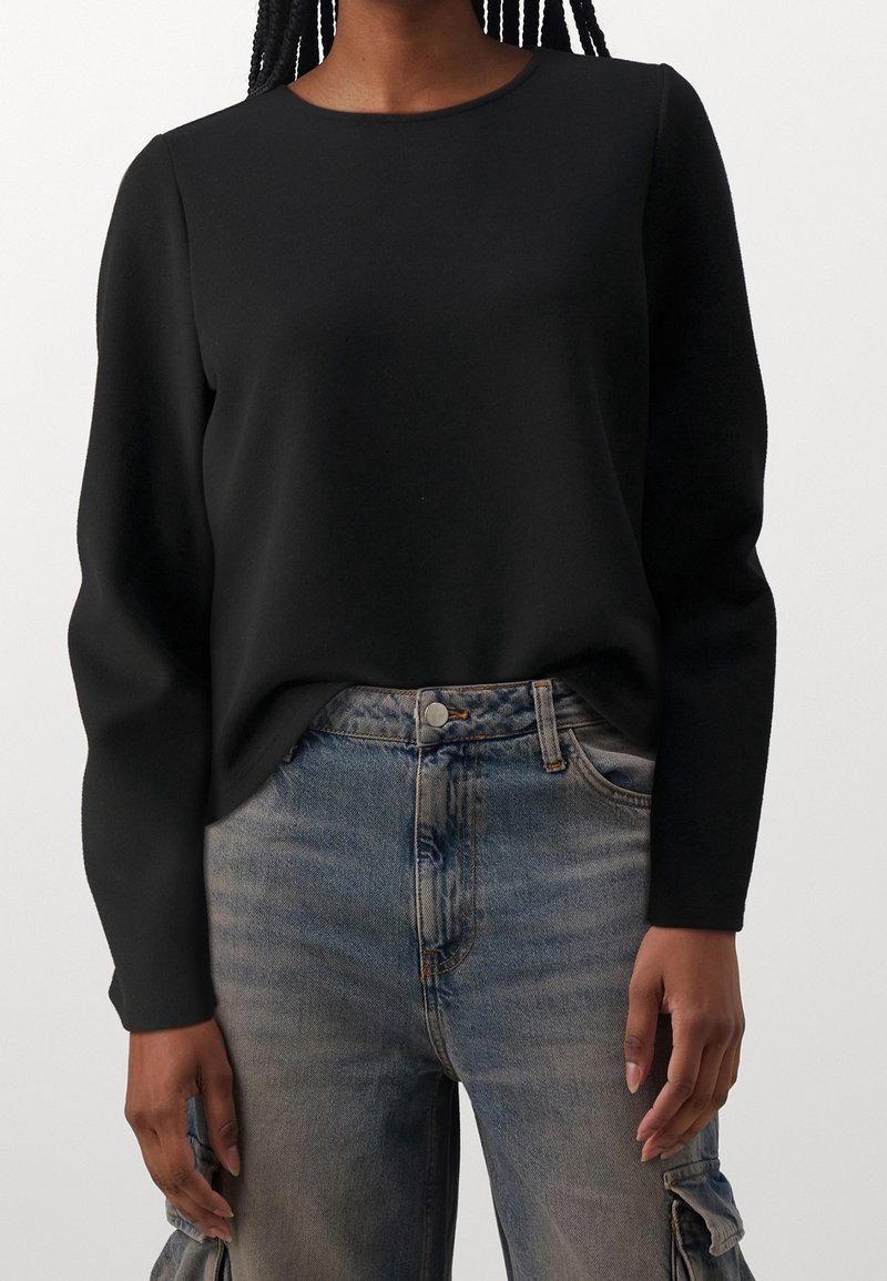 Personne portant une chemise noire à manches longues rentrée dans un jean bleu délavé avec des poches cargo, debout devant un fond uni.