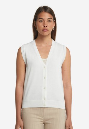 Vit ärmlös stickad väst med V-ringning, framknäppning och ribbkant, bärs över en lätt beige topp.