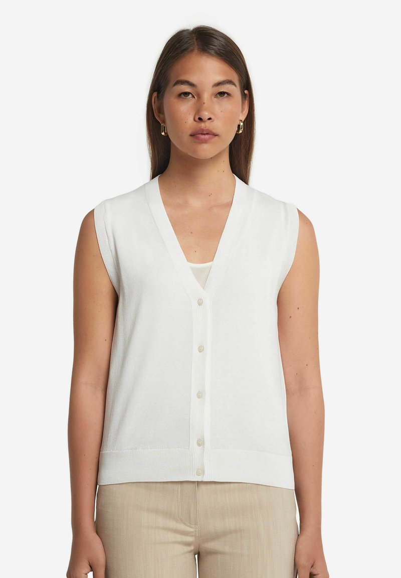 Vit ärmlös stickad väst med V-ringning, framknäppning och ribbkant, bärs över en lätt beige topp.