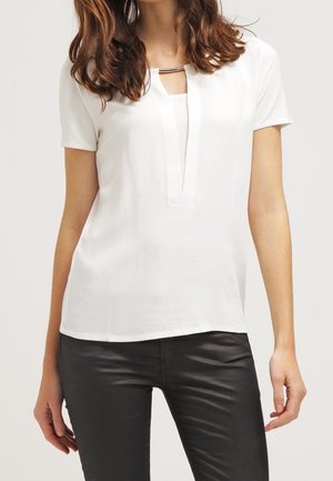 Femme portant un chemisier blanc à manches courtes avec un détail barre métallique au niveau de l'encolure et un pantalon noir ajusté, debout devant un fond uni.