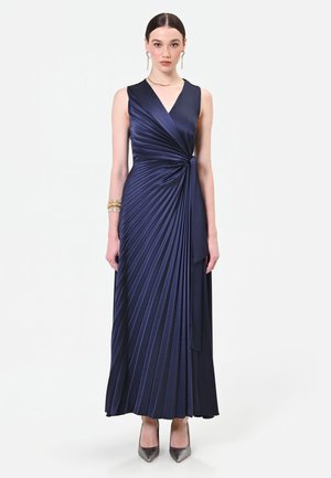 Frau trägt ein ärmelloses, marineblaues, plissiertes Wickelkleid mit V-Ausschnitt und seitlicher Schleife, kombiniert mit metallic spitzen Schuhen und goldenen Schmuck.