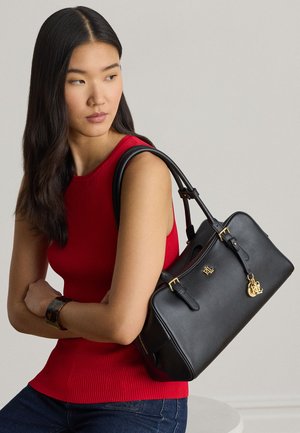 Donna con top rosso senza maniche che indossa una borsa nera in pelle con dettagli dorati sulla spalla, guarda di lato su uno sfondo semplice.