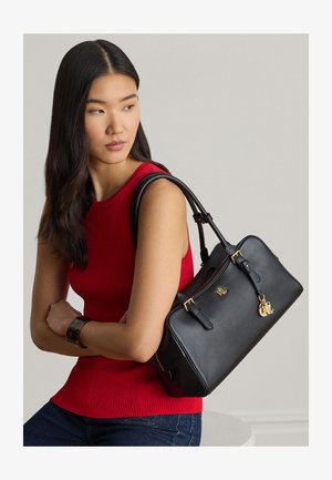 Moteris raudonu be rankovių viršutiniu drabužiu, nešiojanti juodą odinę rankinę su auksinėmis detalėmis ant peties, žiūrinti į šoną prieš vienspalvį foną.