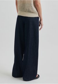 Široke temno modre kavbojke z gladko teksturo in minimalnimi detajli, ujemajoče se z lahkim pletenim zgornjim delom in črnimi sandali.