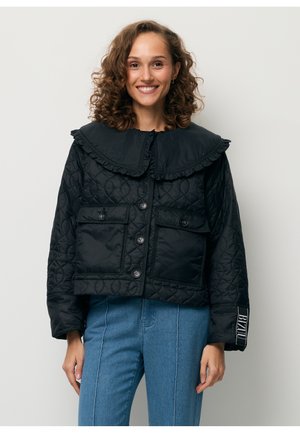 Vrouw met krullend haar, gekleed in een zwarte doorgestikte jas met een grote kraag en een blauwe spijkerbroek, glimlachend tegen een effen lichte achtergrond.