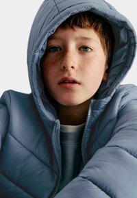 Ung person med fräknar och ljusbrunt hår som bär en zipad ljusblå huva-puffjacka och ser lätt uppåt.