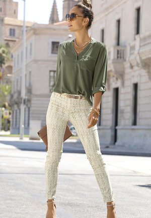 Blouse olive verte à boutons avec des manches retroussées, pantalon skinny crème à motifs, et une pochette marron. Porté avec des accessoires en or et des lunettes de soleil.
