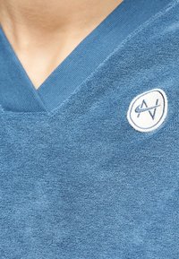 Modra teksturirana majica z v-izrezom in majhnim belim in modrim vezenim okroglim logotipom na prsih.