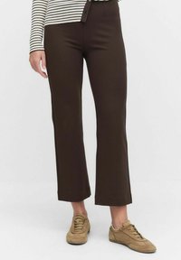 Persoon in donkerbruine rechte broek, beige sneakers en een beige met zwarte gestreepte langemouwtop, staande op een witte achtergrond.