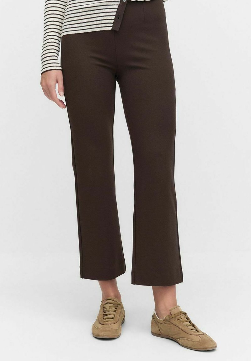 Persoon in donkerbruine rechte broek, beige sneakers en een beige met zwarte gestreepte langemouwtop, staande op een witte achtergrond.