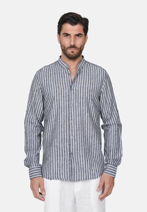 Uomo con capelli scuri e barba che indossa una camicia grigia e bianca a righe verticali con pantaloni bianchi, in piedi su uno sfondo semplice.