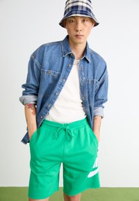 Chemise en jean sur un débardeur blanc, accompagnée de shorts verts à cordon de serrage arborant un logo blanc. Le mannequin porte un chapeau de seau à carreaux.