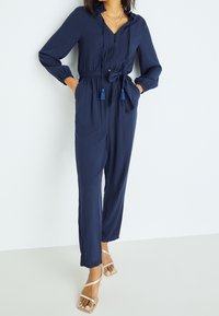 Navy blue jumpsuit in a textured fabric, featuring a button-down front, hood, elastic cuffs, waist tie with tassels, and straight legs.