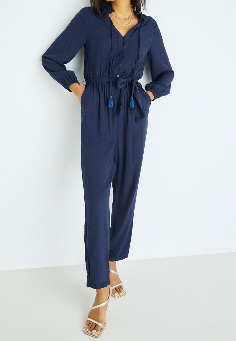 Navy blue jumpsuit in a textured fabric, featuring a button-down front, hood, elastic cuffs, waist tie with tassels, and straight legs.