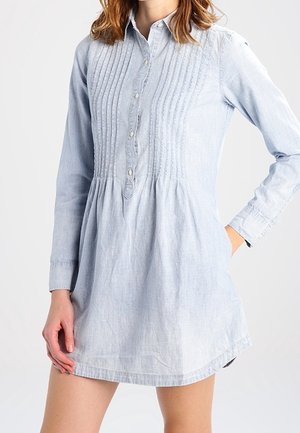 Kobieta ubrana w jasnoniebieską sukienkę-koszulę z długim rękawem, zapiętą na guziki z przodu, z pionowymi plisami na piersi.