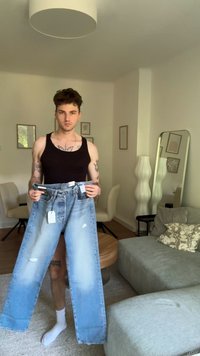 Ung man i svart linne som håller i ljusblå trasiga jeans med prislappar, stående i vardagsrum med soffa, stolar och golvlampa.